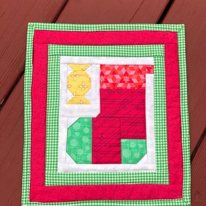 May include: A red and green patchwork quilt block with a stocking design. The stocking is made of red, white, and green fabric with a yellow candy cane.