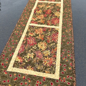May include: A quilted table runner with a floral pattern in shades of red, orange, yellow, and green. The runner has a cream-colored border and a green background with a subtle pattern of red berries.