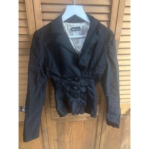 May include: Black blazer with a fitted waist and three decorative buttons. The blazer has long sleeves and a notched lapel. The interior lining is a light beige color, and the label reads "wetseal". The blazer is hanging on a white hanger.