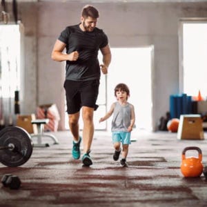 Op de afbeelding: Een persoon in een donkergrijs t-shirt en zwarte sportbroek rent naast een klein kind in een lichtgrijs hemdje en lichtblauwe korte broek in een sportschool met diverse fitnessapparatuur zoals halters en een oranje kettlebell.
