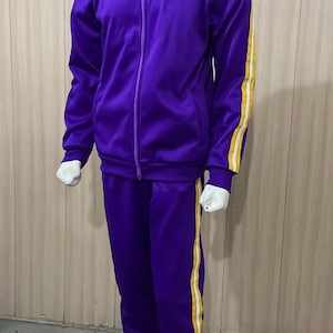 Può includere: Una tuta sportiva viola composta da una giacca con zip e collo alto e pantaloni abbinati. Sia le maniche della giacca che le gambe dei pantaloni presentano una striscia verticale gialla fiancheggiata da due sottili linee bianche laterali.