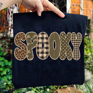 May include: A black t-shirt featuring the word SPOOKY in large, bold letters. Each letter has a distinct pattern: leopard print, camouflage, black and tan buffalo plaid, vertical stripes, polka dots, and a black and tan houndstooth pattern.