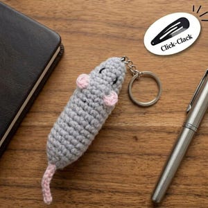 May include: A handmade crochet mouse keychain in gray with pink ears and tail. The mouse has black eyes and is attached to a silver keyring. The image also includes a pen, a notebook, and a sticker that says "Click-Clack".