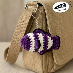 May include: A handmade, purple, white, and black crochet fish keychain hanging from a silver keyring. The fish has white stripes and black outlines. The keychain is attached to a tan bag. The text "Click-Clack" is in the upper right corner.