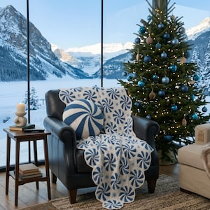 May include: A cozy living room scene with a decorated Christmas tree and a leather armchair. The chair is adorned with a blue and white swirl-patterned throw blanket and a matching round pillow. A small side table holds a candle and books, with a snowy mountain view in the background.