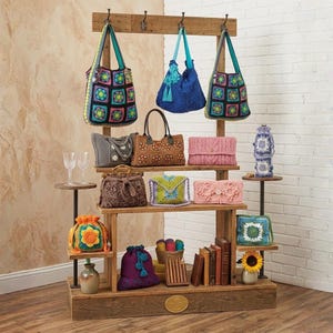 May include: A wooden display stand showcasing a variety of handmade crochet bags and accessories. The bags feature colorful floral and geometric patterns. Other items include a sunflower, books, and yarn.