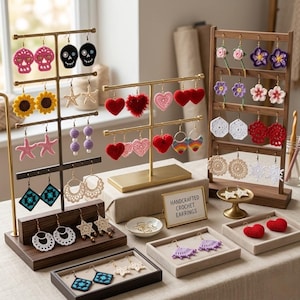 Può includere: Esposizione di orecchini all'uncinetto fatti a mano con vari disegni, tra cui cuori, teschi, fiori e forme geometriche. Gli orecchini sono esposti su supporti dorati e in legno, con un cartello che recita "Handcrafted Crochet Earrings".