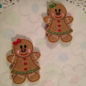 May include: Two felt gingerbread people ornaments with white icing details, red buttons, and green holly accents. The ornaments are on a white doily.