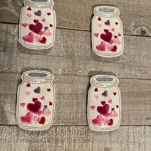 May include: Four white fabric mason jars with pink, red, and purple embroidered hearts inside. The jars are on a wood background.