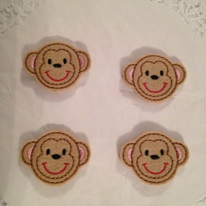 May include: Four brown felt monkey faces with pink ears and red smiling mouths. The monkey faces are embroidered with black eyes and a black nose.
