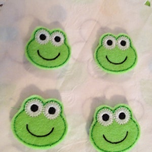May include: Four green felt frog appliques with black eyes and a white outline. Each frog has a black stitched smile.