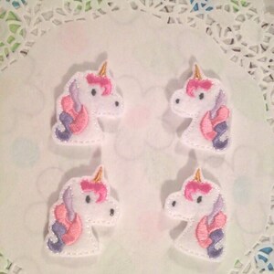 May include: Four white felt unicorn head appliques with pink and purple manes and gold horns. The appliques are on a white doily background.