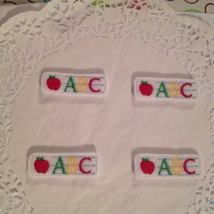 May include: Four white fabric patches with red apple and ABC embroidered in green, yellow, and blue. The patches are on a white doily.