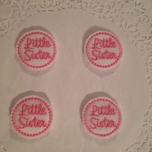 May include: Four white fabric circles with pink stitching around the edges. Each circle has the words "Little Sister" embroidered in pink thread.