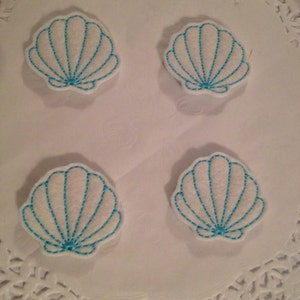 May include: Four white felt seashells with blue embroidery. The seashells are arranged in a square pattern on a white background.