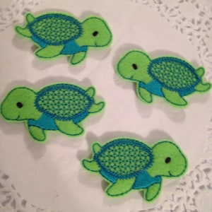 May include: Four green and blue felt turtle appliques with embroidered details. The turtles are on a white doily background.