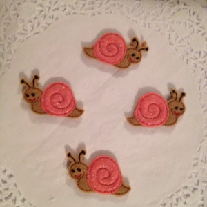 May include: Four felt snail appliques with pink shells and brown bodies. The snails have black eyes and smiling faces.