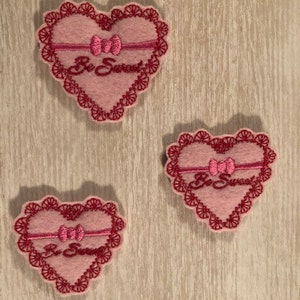 Könnte beinhalten: Drei herzförmige Patches aus rosa Filz mit roten gestickten Rändern. Jeder Patch hat eine rosa Schleife und den Text "Be Sweet" in rot gestickt.