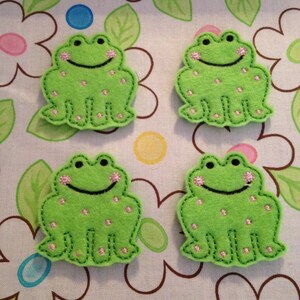 May include: Four green felt frog appliqués with pink accents and black stitched details. Each frog has a smiling face and pink polka dots. They are arranged on a floral patterned fabric with white, pink, and blue flowers.