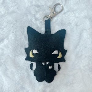 May include: A black felt keyring shaped like a stylised cat character wearing a black waistcoat with white accents. The cat has pointed ears, embroidered yellow and white eyes, and a silver-toned metal lobster clasp attached by a black fabric strap.