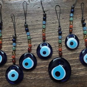 Puede incluir: Siete amuletos de ojo turco de cristal azul oscuro con un diseño de ojo azul claro y blanco. Cada amuleto está ensartado con un cordón negro y cuentas coloridas en tonos de rojo, naranja, amarillo, verde, azul y morado. Los amuletos se muestran sobre una superficie de madera.