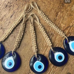 Puede incluir: Varios amuletos de ojo turco de cristal azul con detalles blancos y celeste, cada uno suspendido de un cordón de yute tejido. Los amuletos son redondos y tienen un fondo azul oscuro. Los cordones de yute son de color beige.