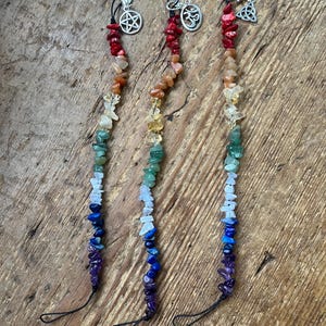 May include: Three chakra stone wands featuring a spectrum of gemstones. Each wand is topped with a silver charm, including a pentagram, an Om symbol, and a Celtic knot. The stones are arranged in a gradient of colours, from red to purple.