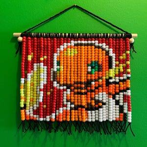 May include: A wall hanging made of colorful plastic pony beads arranged to form a pixelated image of a character with an orange head, green eyes, and yellow accents. The beads are strung on vertical black cords suspended from a wooden dowel rod.