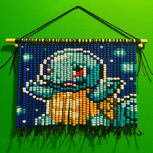 May include: A wall hanging made of colorful plastic beads arranged to form a pixelated image of a blue turtle-like creature with a yellow belly. The beads are strung on black cords suspended from a horizontal wooden dowel with a black hanging cord.