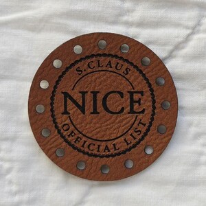 May include: A circular brown leather tag with pre-punched holes around the perimeter. The center features a black stamped design with the text S. CLAUS NICE OFFICIAL LIST arranged in a circular pattern with a decorative border.