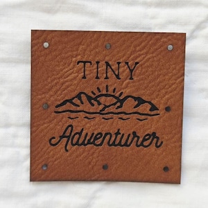 May include: A square brown faux leather patch with a textured surface featuring the black text TINY ADVENTURER and a graphic of mountains and a sun. The patch has six small holes along the edges for attachment, arranged in a rectangular pattern.