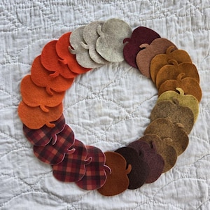 May include: A circular arrangement of felt apple-shaped cutouts in a gradient of colors including cream, orange, brown, and red, with some pieces featuring a red and black plaid pattern, all layered to form a continuous ring shape.