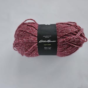 May include: A ball of Cozy Camp yarn by Eddie Bauer. The yarn is a mix of red and white, with a black label wrapped around it. The label states the yarn is 100% recycled acrylic and weighs 3.5 oz (100g).