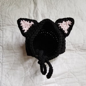 May include: Black crocheted cat hat with pink ears and a tie. The hat is made of soft yarn and has a cute, playful design.