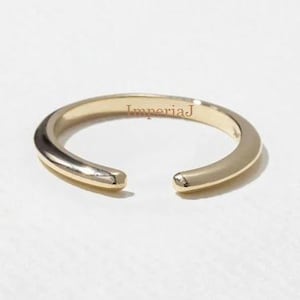 Op de afbeelding: Een goudkleurige open ring met een gladde, gepolijste afwerking. De ring heeft een opening aan de voorkant, waardoor een uniek ontwerp ontstaat. Het woord "ImperiaJ" is op de ring gegraveerd. De ring staat tegen een witte achtergrond.
