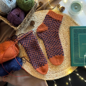 May include: A pair of hand-knitted socks featuring a two-tone geometric pattern in burnt orange and navy blue. One sock is fully completed with a ribbed cuff, while the other remains on circular knitting needles, showing the ongoing work.