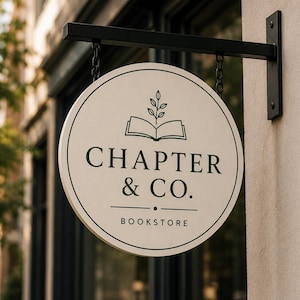 以下が含まれることがあります： 黒い文字で「CHAPTER & CO. BOOKSTORE」と書かれた、丸いクリーム色の本屋の看板。看板は黒い金属製のブラケットから鎖で吊り下げられています。看板には開いた本と葉のグラフィックが描かれています。