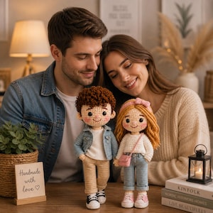May include: Two handmade crochet dolls, a boy and a girl, stand on a wooden surface. The boy doll has brown hair, a blue jacket, and beige pants. The girl doll has blonde hair, a pink bow, and blue jeans. A sign reads "Handmade with love".