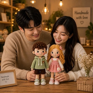 May include: Two handmade crochet dolls, a boy and a girl, are held by a couple. The boy doll wears a green hoodie and brown shorts. The girl doll is in a pink dress. The dolls are on a wooden table. A framed sign reads "Handmade with love".