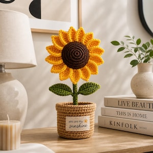 Puede incluir: Un girasol tejido a ganchillo en una maceta tejida. El girasol tiene pétalos amarillos y un centro marrón. La maceta es de color beige con una etiqueta que dice "you are my sunshine". Las hojas son verdes.