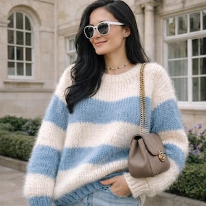 Könnte beinhalten: Ein gemütlicher, übergroßer Pullover mit horizontalen Streifen in Hellblau und Creme. Dazu eine helle Jeans und eine kleine, taupefarbene Handtasche mit goldener Kette. Die Person trägt eine weiße Sonnenbrille. Der Pullover ist ein Blickfang.