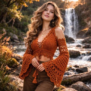 May include: An orange crochet top with long, flared sleeves and a lace-up front. The top has a V-neck and is made of a textured yarn. The sleeves have an openwork design. The top is paired with brown pants. The background features a waterfall and foliage.