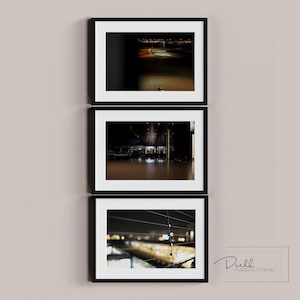 以下が含まれることがあります： 黒いフレームに入った3つのアートプリントが、明るいベージュの壁に展示されています。プリントは、ぼやけた光と暗い背景を持つ夜のシーンを特徴としています。壁の右下隅には「Dull Margin Frame」という文字があります。