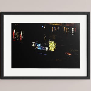 May include: A framed art print featuring a nighttime scene with colorful reflections on water. The image is dominated by dark tones, with vibrant splashes of red, yellow, and blue light. The artwork is encased in a black frame with a white mat.