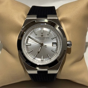 May include: A silver Vacheron Constantin watch with a black rubber strap. The watch face is silver with silver hands and markers. The watch has a date window at the 3 o'clock position. The watch is displayed on a beige cushion.