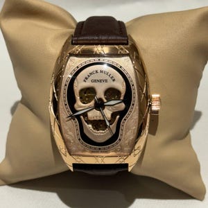 May include: A gold-toned wristwatch with a brown leather band. The watch face features a skull design with the words "FRANCK MULLER GENEVE" and "SWISS MADE". The watch is displayed on a beige pillow.