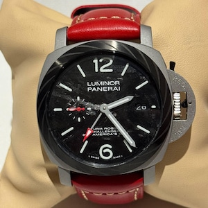 May include: A close-up of a Panerai Luminor watch with a black face, white numerals, and a date display. The watch has a grey case and a red leather strap. The watch face has the text "LUMINOR PANERAI".