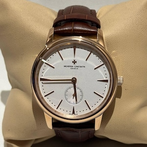 May include: A Vacheron Constantin Geneve wristwatch with a round rose gold case, a silver dial featuring thin gold hour markers, a small seconds sub-dial at the six o'clock position, and a dark brown leather strap with a textured finish.