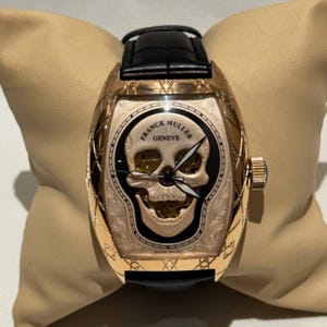 Könnte beinhalten: Eine Luxus-Armbanduhr mit einem goldfarbenen Gehäuse und einem schwarzen Lederarmband. Das Zifferblatt der Uhr zeigt ein Totenkopf-Design mit den Worten "FRANCK MULLER GENEVE". Die Uhr wird auf einem beigen Kissen präsentiert.