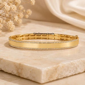 May include: A gold bangle bracelet featuring a smooth, polished central band bordered by two rows of small, clear pavé-set stones. The bracelet has a hinged design with a visible clasp mechanism and the text EliteDiamoraJewels printed above it.